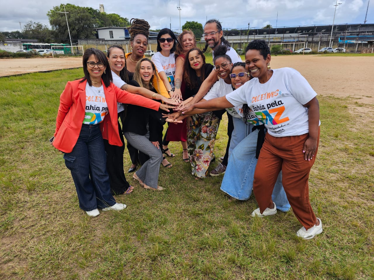 Grupo de pessoas em um terreno, ao ar livre, juntando as mãos em símbolo de parceria.