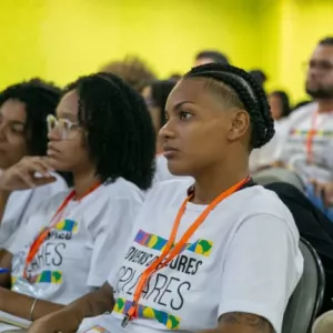 Jovens negras, vestindo a camiseta de Jovens Defensores Populares, sentadas como se ouvissem uma palestra.