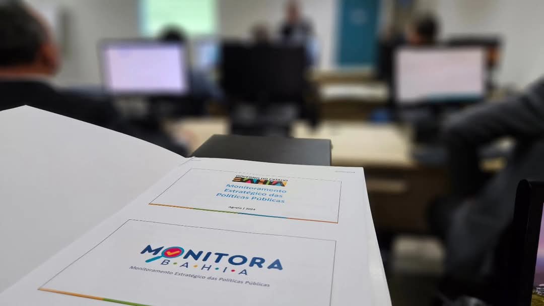 imagem da marca do Monitora no papel sobre a mesa. Fundo desfocado de uma reunião