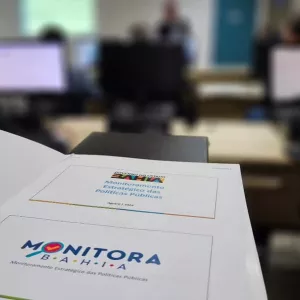 imagem da marca do Monitora no papel sobre a mesa. Fundo desfocado de uma reunião