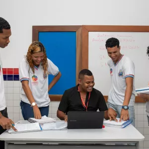 Foto ilustrativa de professsor sentado e quarto alunos em volta. Todos sorrindo