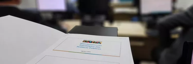 imagem da marca do Monitora no papel sobre a mesa. Fundo desfocado de uma reunião