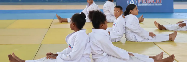 Crianças sentadas no tatame com total de 6 crianças e a cada par uma de costas para outra