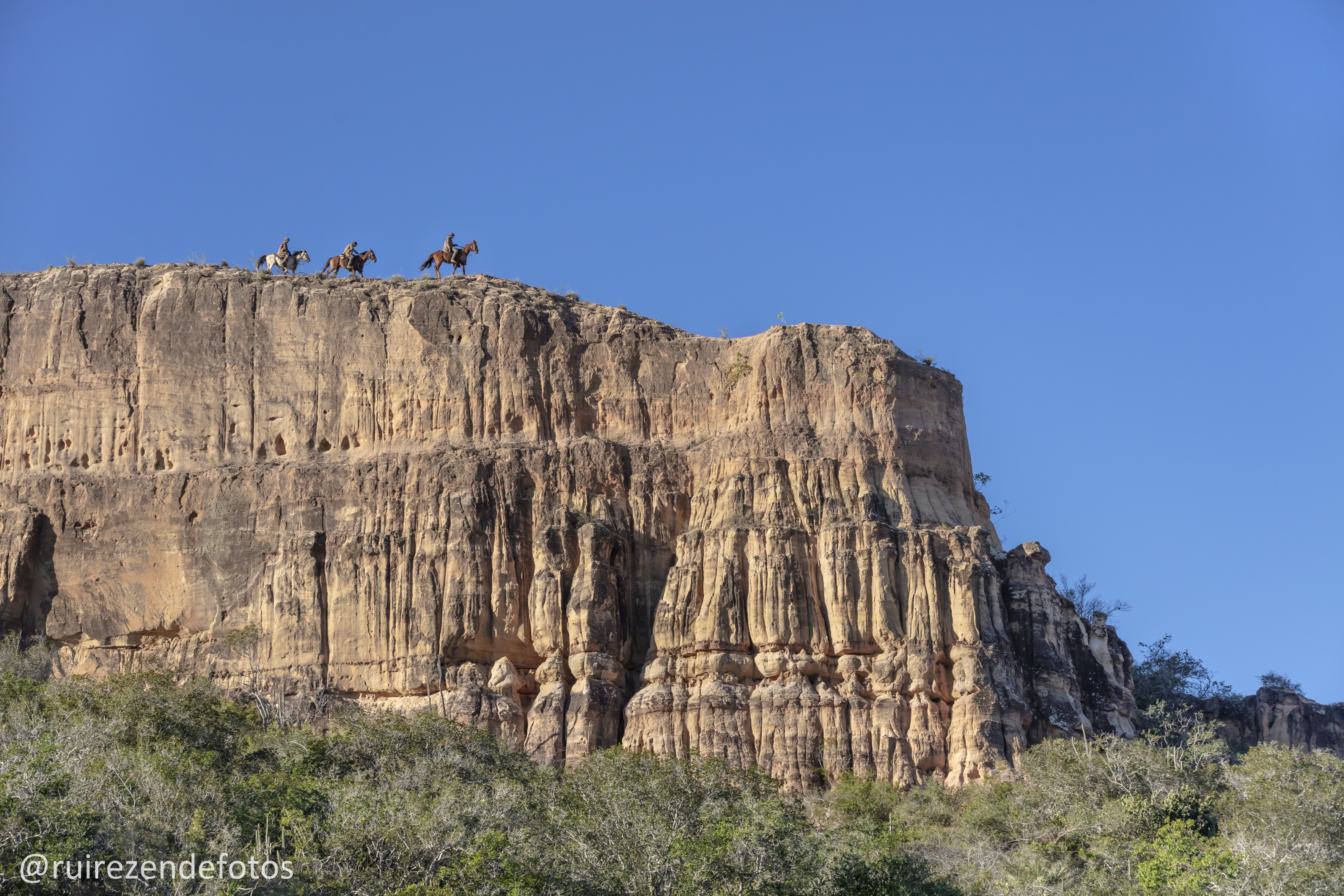 vaqueiros ao longe, montados em cavalos, em cima de um imenso paredão. Céu azul