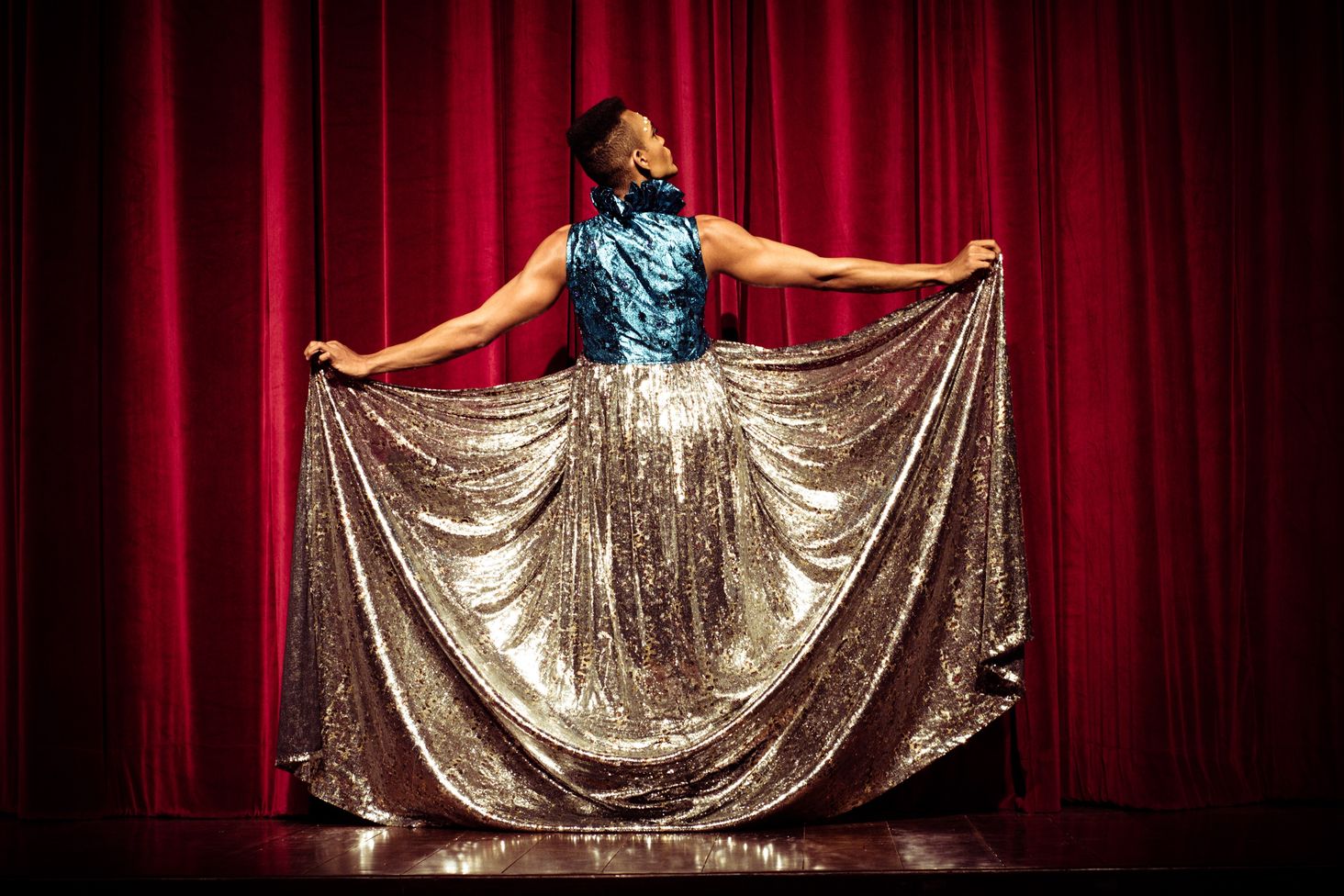 Homem de costas com vestido brilhante. Parte de cima azul e parte da saia dourada. No fundo cortinas vermelhas
