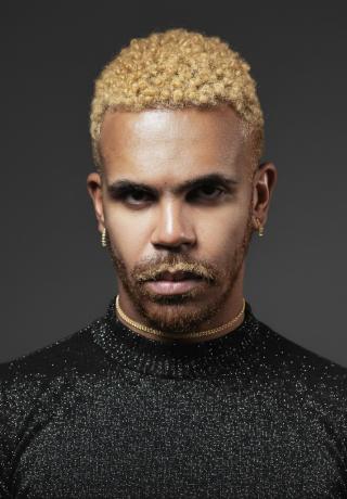 Foto com homem negro, cabelo loiro claro, veste uma camisaa de manga cor preto. 