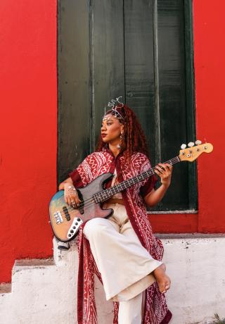 Mulher negra, veste calça branca folgada, camisa longa vermelha, cabelos vermelhos. Ela está sentada apoiada em uma escada cor branca segurando um violão no colo, em frente a fachada de uma casa. As paredes da fachada é vermelha e porta alta cor verde. 