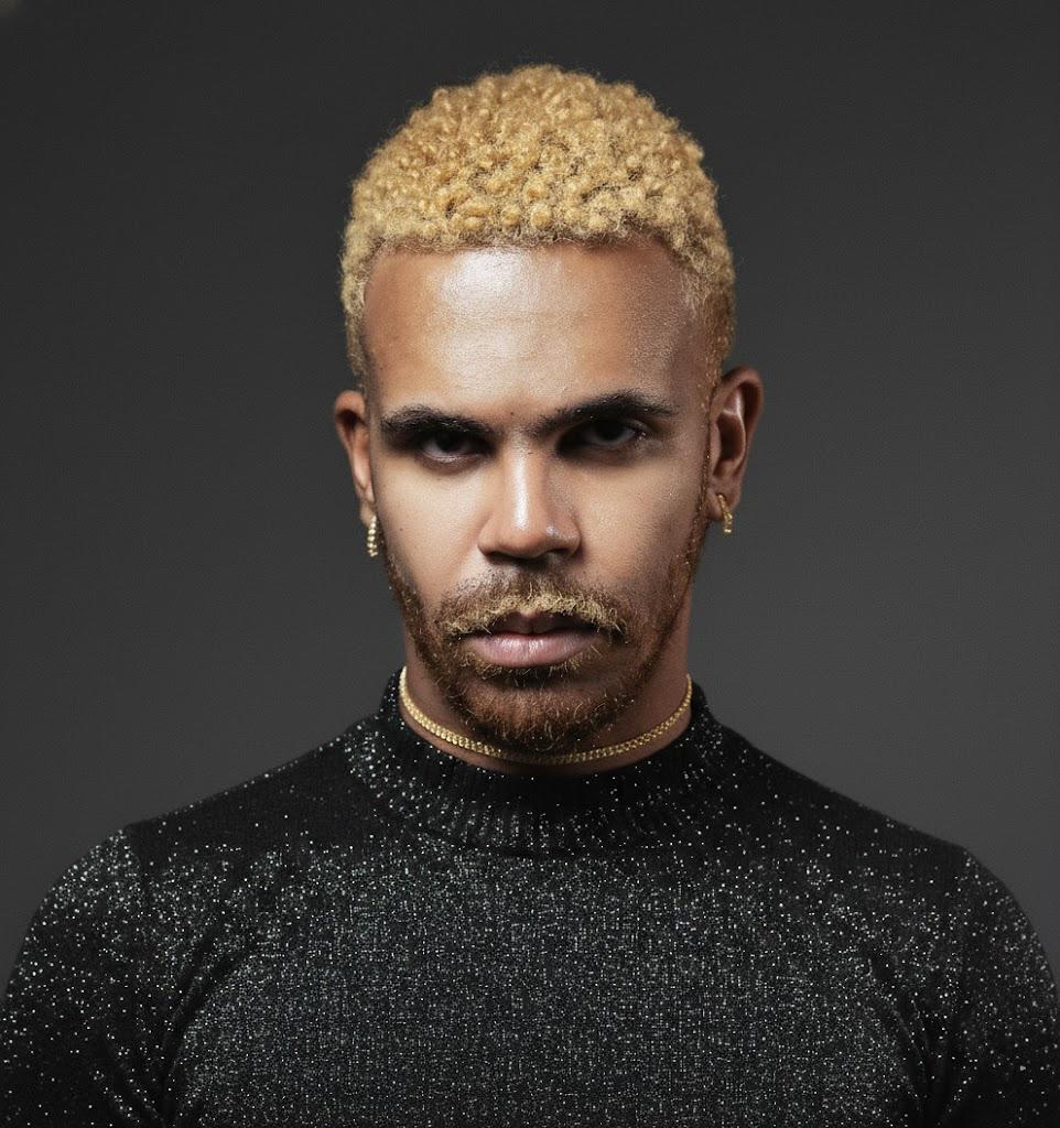 Foto com homem negro, cabelo loiro claro, veste uma camisaa de manga cor preto. 