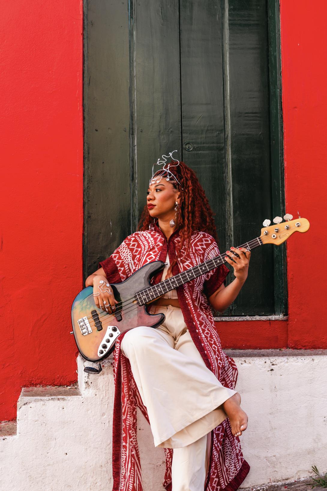 Mulher negra, veste calça branca folgada, camisa longa vermelha, cabelos vermelhos. Ela está sentada apoiada em uma escada cor branca segurando um violão no colo, em frente a fachada de uma casa. As paredes da fachada é vermelha e porta alta cor verde. 