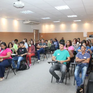 Um grupo de cerca de 30 pessoas está sentado em uma sala de aula, prestando atenção ao que acontece na frente. A sala é bem iluminada, com painéis de madeira e um projetor no teto. O público é diverso e parece concentrado, com alguns fazendo anotações ou usando seus celulares.