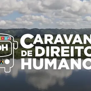  A imagem mostra o logotipo da "Caravana de Direitos Humanos" em destaque, com a sigla "SJDH" (Secretaria de Justiça e Direitos Humanos) dentro de um ícone de ônibus estilizado, representando o projeto itinerante. O fundo exibe uma paisagem natural, com um rio ou lago refletindo o céu azul e nuvens, cercado por vegetação densa e montanhas ao longe.