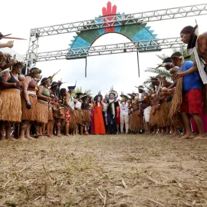 Na imagem, um grupo de indígenas forma um corredor humano em trajes tradicionais, com saias de palha, cocares e pinturas corporais. No centro, lideranças ou convidados especiais, incluindo uma mulher de vestido vermelho, se destacam sob uma estrutura decorativa colorida ao fundo. A cena transmite um clima de solenidade e respeito cultural.