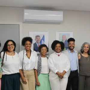 Na imagem, um grupo de oito pessoas está alinhado lado a lado, sorrindo para a câmera. O fundo exibe paredes claras e quadros de autoridades políticas ao centro.