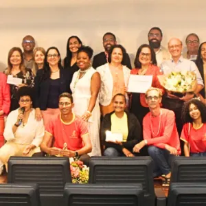 A imagem mostra um grupo de pessoas em um evento de premiação ou reconhecimento. Estão reunidos em um ambiente interno, com cadeiras de auditório ao fundo. As pessoas, tanto homens quanto mulheres, variam em idade e aparência, algumas segurando certificados e flores. Há uma atmosfera de celebração e reconhecimento, com sorrisos e expressões de satisfação. Algumas pessoas estão em pé e outras estão sentadas, criando uma disposição dinâmica e informal no espaço.