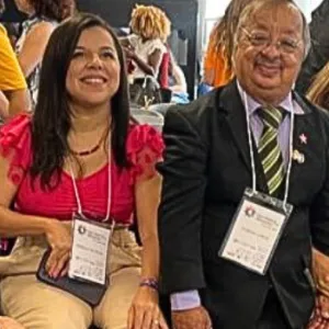 A imagem mostra um grupo de cinco pessoas com deficiência reunidas em um evento. Elas estão sentadas, algumas em cadeiras de rodas, sorrindo e usando crachás pendurados no pescoço. Ao fundo, há outras pessoas em pé e movimentando-se, sugerindo um ambiente de confraternização ou conferência. As pessoas estão em trajes formais ou casuais, com expressões acolhedoras e animadas. A cena transmite inclusão e participação ativa em um espaço social ou institucional.