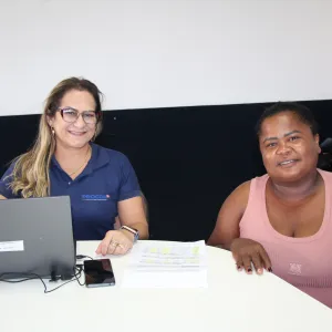 Duas mulheres estão sentadas em uma mesa durante um atendimento no mutirão de renegociação de dívidas do Procon-BA; uma delas, vestindo uma camisa do órgão, sorri ao lado da consumidora, que também sorri e tem documentos sobre a mesa.