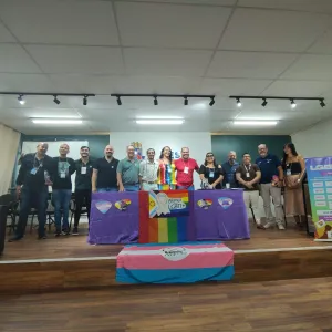 A imagem mostra um grupo diverso de pessoas reunidas em um auditório, posando para a foto atrás de uma mesa coberta com bandeiras do movimento LGBTQIA+. No centro, há um troféu colorido com a inscrição "Aliança LGBTI+". À direita, um banner exibe a programação de um evento voltado para a comunidade LGBTQIA+ em Jequié, Bahia. A cena transmite um clima de celebração, engajamento e defesa dos direitos da população LGBTQIA+, reforçando a importância do diálogo e da construção de políticas inclusivas.