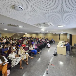 Auditório cheio com pessoas da plateia sentadas de frente para o palestrante Paulo Miguez. O palestrante está sentado à mesa, ao fundo, um painel virtual com o tema da palestra “A importância socioeconômica e cultural do Carnaval para a Bahia”.  