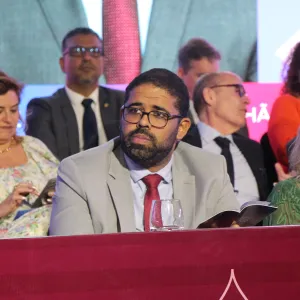 A imagem mostra um homem negro de barba e óculos, vestindo um terno cinza e gravata vermelha, sentado em uma mesa coberta com um tecido bordô. Ele segura um livro e tem uma expressão atenta. Ao seu redor, há várias pessoas bem-vestidas, algumas em trajes formais, outras em roupas estampadas. No fundo, há um painel colorido com letras parcialmente visíveis e pessoas conversando. A cena parece ser de um evento formal ou uma cerimônia.