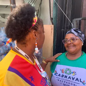  Duas mulheres conversam amigavelmente. Uma delas veste uma camisa verde com a estampa "CARNAVAL SOLIDÁRIO SALVADOR 2025" e parece sorridente. Ao fundo, há várias garrafas plásticas empilhadas, indicando um possível esforço de reciclagem durante um evento.  Se precisar de algo mais, é só falar! 😊