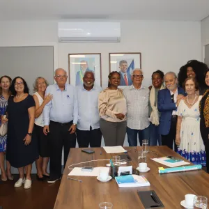 : Um grupo de pessoas reunido para uma foto ao final da reunião. Eles estão sorrindo e posando juntos, demonstrando um momento de integração e encerramento do encontro. A mesa ainda contém materiais da reunião, como cadernos, canetas e xícaras de café. Ao fundo, há dois retratos de figuras políticas na parede.