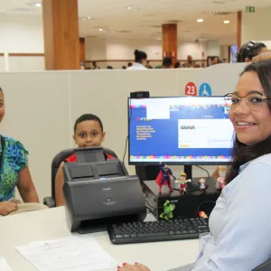 Na imagem, vemos um ambiente interno de atendimento ao público, possivelmente em um órgão governamental. À esquerda, uma mulher está sentada sorrindo, acompanhada de um menino ao seu lado. Ambos estão sendo atendidos por uma funcionária que está do outro lado da mesa, sorridente e voltada para a câmera. A funcionária usa óculos e uma camisa azul clara, e está sentada em frente a um computador com a tela exibindo informações do sistema do Governo da Bahia, mais especificamente sobre o CIPTEA.