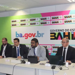A imagem mostra uma reunião institucional com representantes do Governo do Estado da Bahia, no Brasil. O fundo exibe repetidamente o logotipo do governo e a URL "ba.gov.br", indicando um evento oficial. Há seis pessoas sentadas à mesa, com notebooks e microfones à frente, sugerindo uma apresentação formal ou conferência.