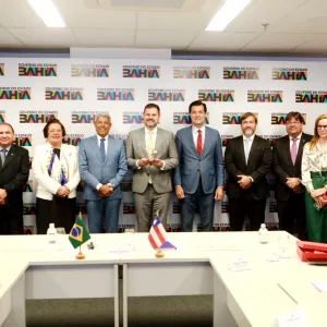 A imagem mostra um grupo de 12 pessoas (8 homens e 4 mulheres) posando formalmente para uma foto em um ambiente institucional. Ao fundo, há um painel com a logomarca do "Governo do Estado da Bahia". No centro da mesa, há duas bandeirinhas — uma do Brasil e outra da Bahia — indicando um evento oficial. A pessoa no centro segura um troféu ou prêmio, sugerindo que se trata de uma cerimônia de reconhecimento ou premiação. Todos estão trajando roupas formais, reforçando o caráter oficial da ocasião.