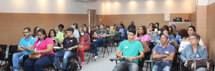 Um grupo de cerca de 30 pessoas está sentado em uma sala de aula, prestando atenção ao que acontece na frente. A sala é bem iluminada, com painéis de madeira e um projetor no teto. O público é diverso e parece concentrado, com alguns fazendo anotações ou usando seus celulares.