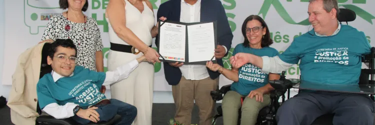A imagem mostra cinco pessoas sorrindo em um evento da "Caravana de Direitos Humanos" durante o "Setembro Verde". Duas estão em cadeiras de rodas, usando camisetas azuis com o slogan "Acesso à Justiça e Promoção de Direitos". No centro, uma mulher e um homem seguram um documento em destaque, enquanto um arranjo de flores está à frente do grupo. A foto transmite uma celebração dos direitos humanos e da inclusão de pessoas com deficiência.