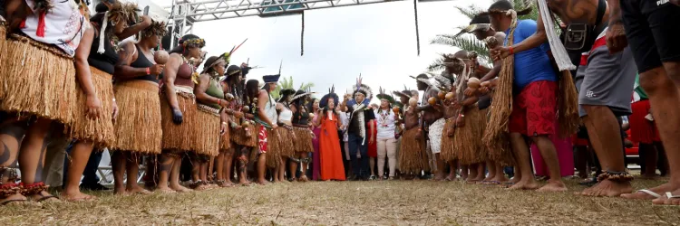 Na imagem, um grupo de indígenas forma um corredor humano em trajes tradicionais, com saias de palha, cocares e pinturas corporais. No centro, lideranças ou convidados especiais, incluindo uma mulher de vestido vermelho, se destacam sob uma estrutura decorativa colorida ao fundo. A cena transmite um clima de solenidade e respeito cultural.