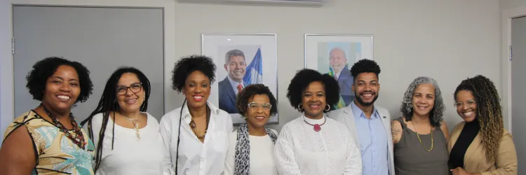 Na imagem, um grupo de oito pessoas está alinhado lado a lado, sorrindo para a câmera. O fundo exibe paredes claras e quadros de autoridades políticas ao centro.