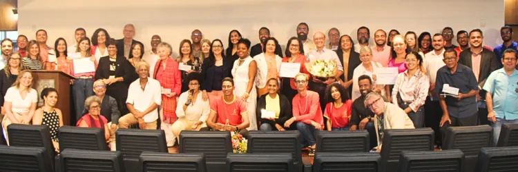 A imagem mostra um grupo de pessoas em um evento de premiação ou reconhecimento. Estão reunidos em um ambiente interno, com cadeiras de auditório ao fundo. As pessoas, tanto homens quanto mulheres, variam em idade e aparência, algumas segurando certificados e flores. Há uma atmosfera de celebração e reconhecimento, com sorrisos e expressões de satisfação. Algumas pessoas estão em pé e outras estão sentadas, criando uma disposição dinâmica e informal no espaço.