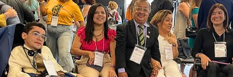 A imagem mostra um grupo de cinco pessoas com deficiência reunidas em um evento. Elas estão sentadas, algumas em cadeiras de rodas, sorrindo e usando crachás pendurados no pescoço. Ao fundo, há outras pessoas em pé e movimentando-se, sugerindo um ambiente de confraternização ou conferência. As pessoas estão em trajes formais ou casuais, com expressões acolhedoras e animadas. A cena transmite inclusão e participação ativa em um espaço social ou institucional.