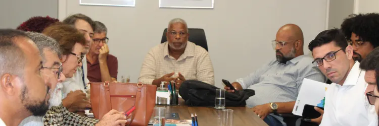 Uma sala de reuniões com uma grande mesa retangular, onde várias pessoas estão reunidas em um encontro estratégico. Algumas estão tomando notas, outras usam laptops e dispositivos móveis, enquanto uma mulher ao fundo, próxima a uma tela de apresentação, conduz a discussão. A tela exibe logotipos de diferentes organizações e a palavra "parceiros". O ambiente é moderno e bem iluminado, com quadros decorativos de arte indígena nas paredes. A diversidade dos participantes reflete um grupo engajado em planejamen