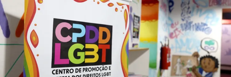 A imagem mostra um banner do Centro de Promoção e Defesa dos Direitos LGBT (CPDD LGBT). O banner tem um fundo branco com bordas coloridas em tons do arco-íris e o nome do centro escrito em letras grandes e coloridas. Abaixo do nome, há um QR code.