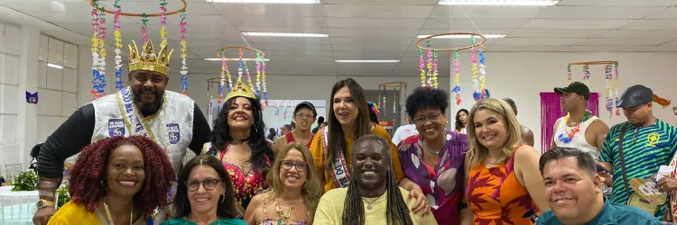 Pessoas reunidas, de roupas coloridas, num cenário carnavalesco. 