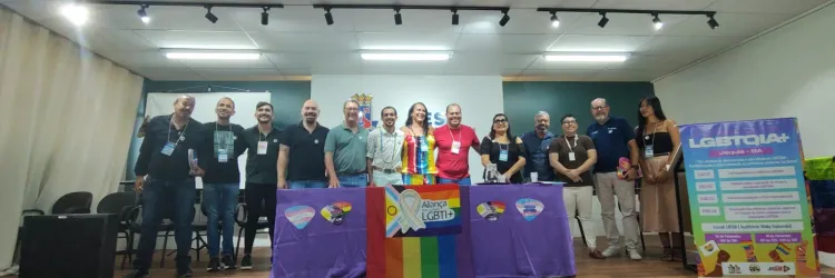 A imagem mostra um grupo diverso de pessoas reunidas em um auditório, posando para a foto atrás de uma mesa coberta com bandeiras do movimento LGBTQIA+. No centro, há um troféu colorido com a inscrição "Aliança LGBTI+". À direita, um banner exibe a programação de um evento voltado para a comunidade LGBTQIA+ em Jequié, Bahia. A cena transmite um clima de celebração, engajamento e defesa dos direitos da população LGBTQIA+, reforçando a importância do diálogo e da construção de políticas inclusivas.