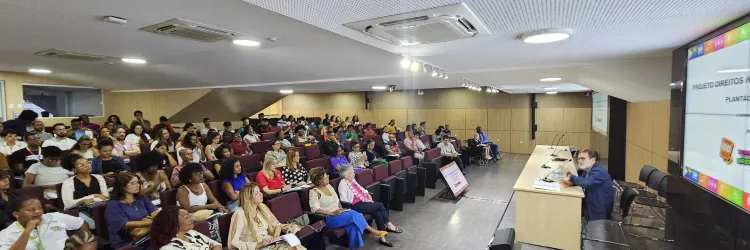 Auditório cheio com pessoas da plateia sentadas de frente para o palestrante Paulo Miguez. O palestrante está sentado à mesa, ao fundo, um painel virtual com o tema da palestra “A importância socioeconômica e cultural do Carnaval para a Bahia”.  