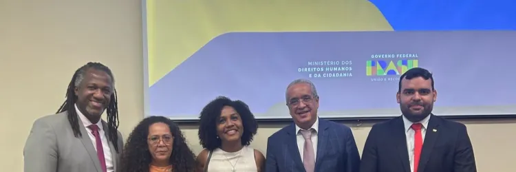 cinco pessoas posam para a câmera em um evento institucional. Elas estão vestidas de forma formal e sorriem. Atrás delas, há um telão com os logotipos do Ministério dos Direitos Humanos e da Cidadania e do Governo Federal (IDAI - União e Reconstrução).