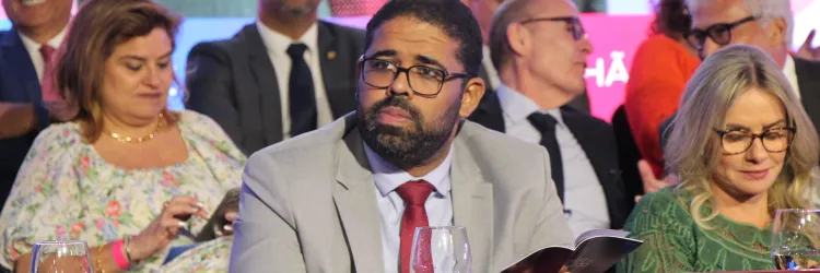 A imagem mostra um homem negro de barba e óculos, vestindo um terno cinza e gravata vermelha, sentado em uma mesa coberta com um tecido bordô. Ele segura um livro e tem uma expressão atenta. Ao seu redor, há várias pessoas bem-vestidas, algumas em trajes formais, outras em roupas estampadas. No fundo, há um painel colorido com letras parcialmente visíveis e pessoas conversando. A cena parece ser de um evento formal ou uma cerimônia.