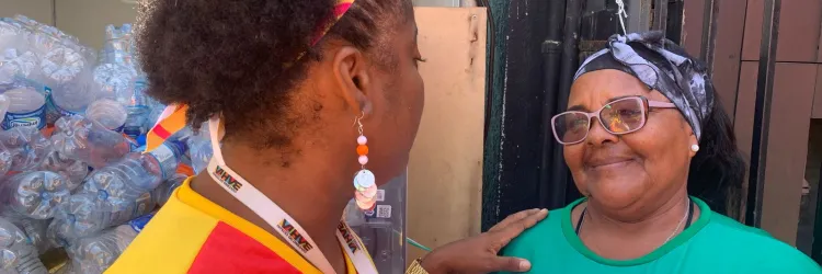  Duas mulheres conversam amigavelmente. Uma delas veste uma camisa verde com a estampa "CARNAVAL SOLIDÁRIO SALVADOR 2025" e parece sorridente. Ao fundo, há várias garrafas plásticas empilhadas, indicando um possível esforço de reciclagem durante um evento.  Se precisar de algo mais, é só falar! 😊