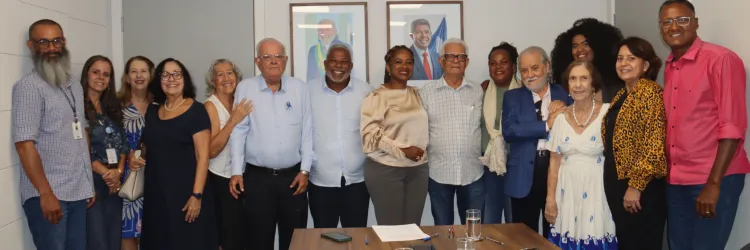 : Um grupo de pessoas reunido para uma foto ao final da reunião. Eles estão sorrindo e posando juntos, demonstrando um momento de integração e encerramento do encontro. A mesa ainda contém materiais da reunião, como cadernos, canetas e xícaras de café. Ao fundo, há dois retratos de figuras políticas na parede.