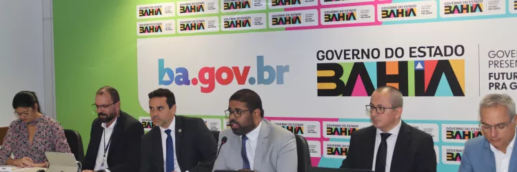A imagem mostra uma reunião institucional com representantes do Governo do Estado da Bahia, no Brasil. O fundo exibe repetidamente o logotipo do governo e a URL "ba.gov.br", indicando um evento oficial. Há seis pessoas sentadas à mesa, com notebooks e microfones à frente, sugerindo uma apresentação formal ou conferência.