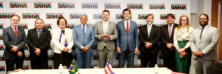 A imagem mostra um grupo de 12 pessoas (8 homens e 4 mulheres) posando formalmente para uma foto em um ambiente institucional. Ao fundo, há um painel com a logomarca do "Governo do Estado da Bahia". No centro da mesa, há duas bandeirinhas — uma do Brasil e outra da Bahia — indicando um evento oficial. A pessoa no centro segura um troféu ou prêmio, sugerindo que se trata de uma cerimônia de reconhecimento ou premiação. Todos estão trajando roupas formais, reforçando o caráter oficial da ocasião.