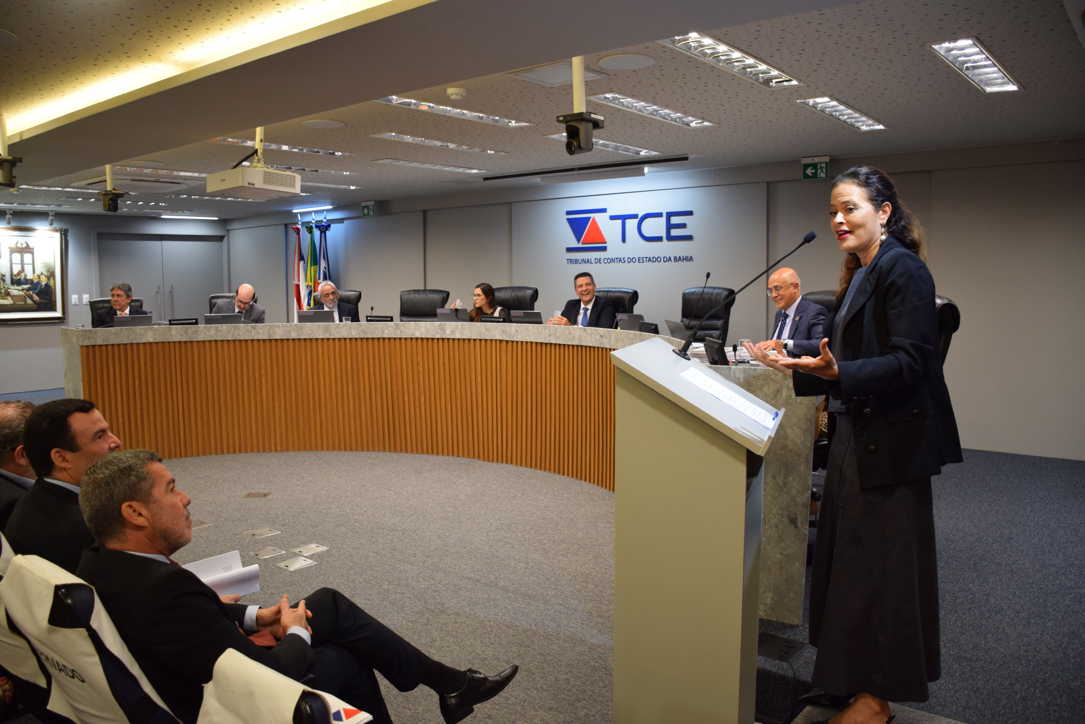Imagem mostra uma mulher em pé falando ao microfone, posicionada atrás de um púlpito no plenário do Tribunal de Contas do Estado da Bahia (TCE-BA). Ao fundo, sete pessoas estão sentadas em uma bancada semicircular, com laptops e microfones à frente. Todos estão com trajes formais. Três homens sentados na plateia observam a oradora com atenção. Na parede, há o logotipo do TCE e bandeiras ao fundo. O ambiente é bem iluminado, moderno e com clima institucional.