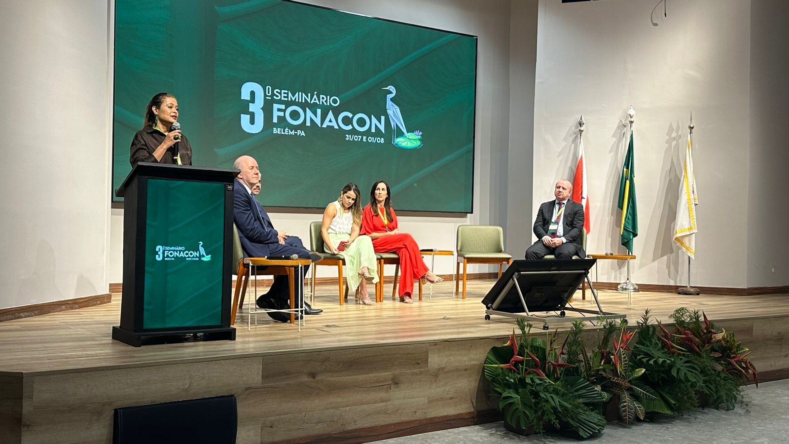 Mulher fala ao microfone em pé, atrás de um púlpito, no palco de um auditório. Ao fundo, uma grande tela exibe “3º Seminário FONACON – Belém-PA – 31/07 e 01/08”. Sentados ao lado da palestrante, há quatro pessoas: três mulheres e dois homens, todos atentos ao evento. À direita do palco, estão três bandeiras: do Pará, do Brasil e uma branca institucional. Na frente do palco, há plantas decorativas com folhagens verdes e flores vermelhas. O ambiente é iluminado, formal e organizado.