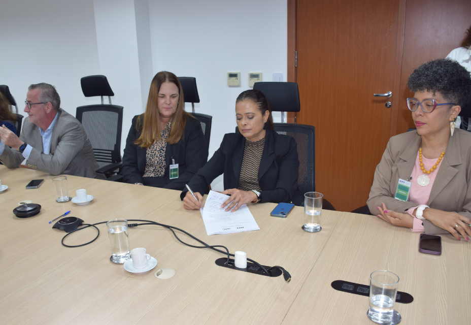 A imagem mostra um grupo de quatro pessoas em uma reunião ao redor de uma mesa. À esquerda, há um homem com cabelo grisalho, usando um terno cinza claro. Ao lado dele, uma mulher com cabelo longo e loiro, vestindo um blazer preto e blusa de estampa. No centro, uma mulher com cabelo preso, usando um terno preto, assina um documento. À direita, uma mulher de cabelo curto e grisalho, com óculos e acessórios coloridos, observa.