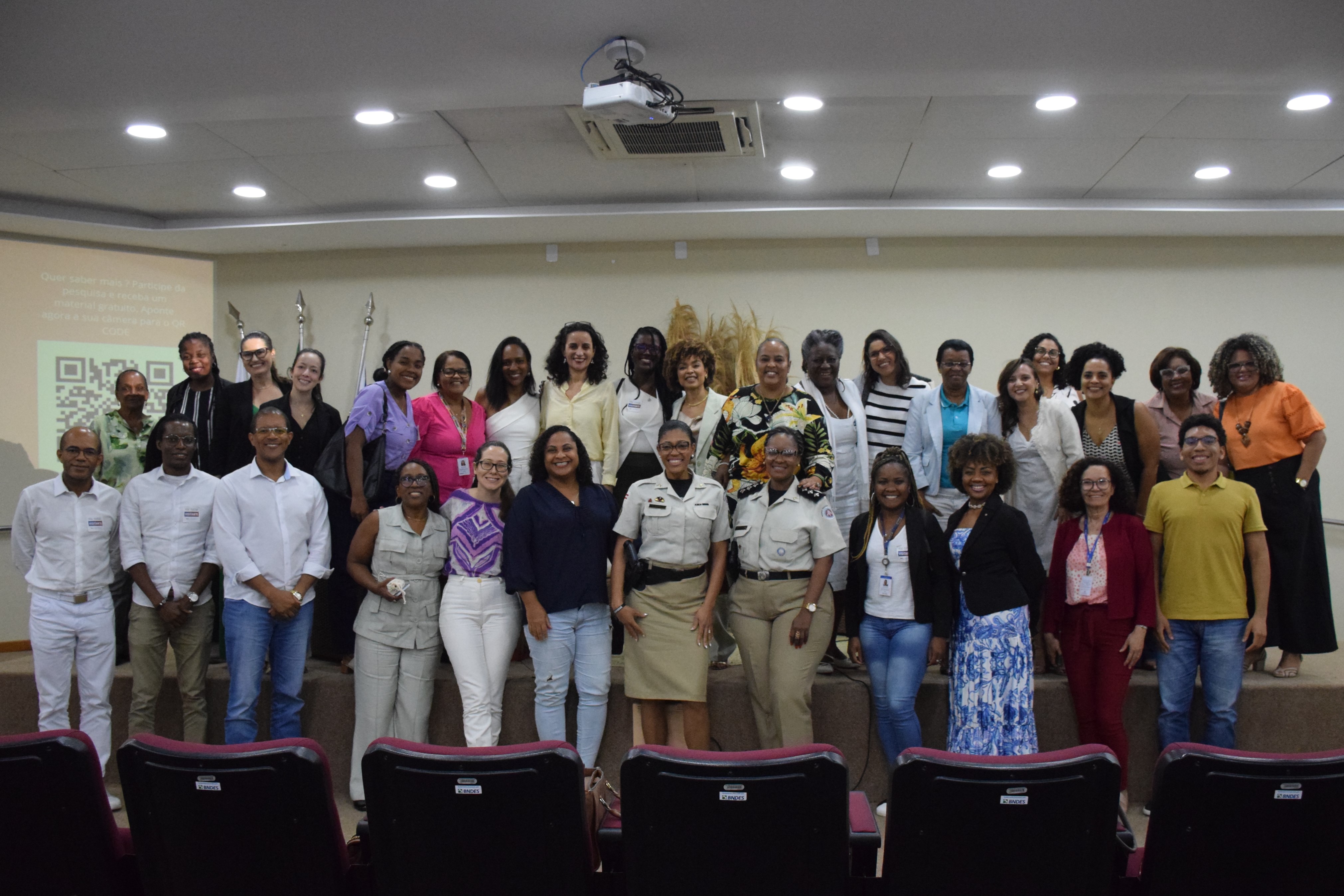 Um grupo diverso de pessoas posa sorrindo para uma foto em um auditório. Elas estão em pé sobre o palco e no chão à frente dele. A maioria são mulheres negras, duas usando uniformes, outras roupas sociais ou casuais. Ao fundo, há bandeiras e um arranjo decorativo com plantas secas. As cadeiras vinho do auditório aparecem na frente da imagem. A foto transmite um clima de encerramento de evento.