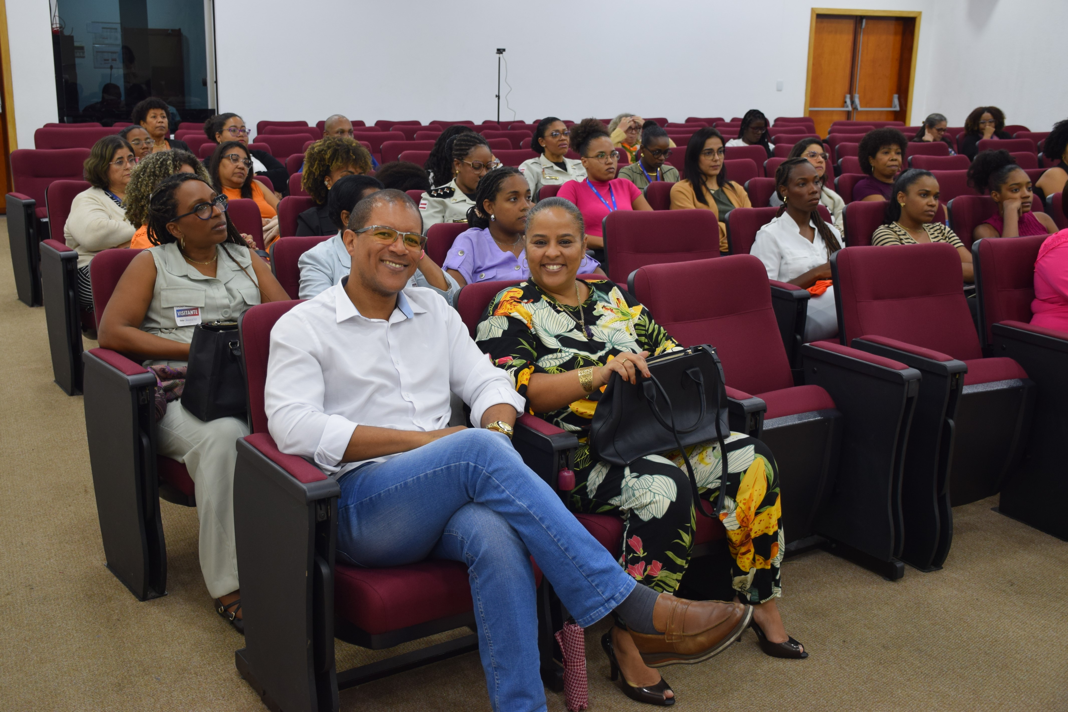 A imagem mostra pessoas sentadas em um auditório com poltronas estofadas na cor vinho. Na frente, um homem de camisa branca e calça jeans está sorrindo, com as pernas cruzadas. Ao lado dele, uma mulher com roupa estampada em tons de preto, amarelo e verde também sorri. Ela segura uma bolsa preta. Ao fundo, outras pessoas, majoritariamente mulheres negras, assistem ao evento com atenção. O ambiente é fechado, bem iluminado e com portas de madeira ao fundo.